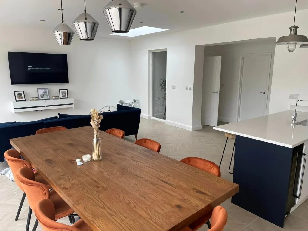 Timber dining table with orange chairs beside a navy sofa and new kitchen