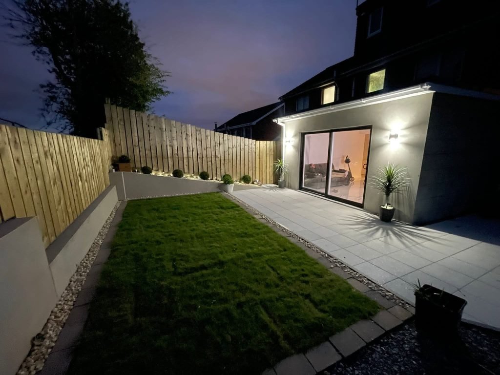 Illuminated extension beside a lawn, pale paving and timber garden fence