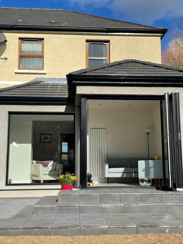 Grey rendered pitched roof extension with dark glazed doors folded open onto the paved terrace